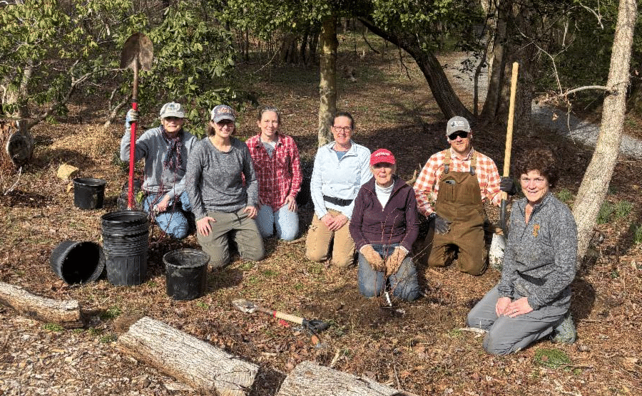 McCoy Garden Group volunteers kneeling together in a wooded garden bed with shovels and plant pots