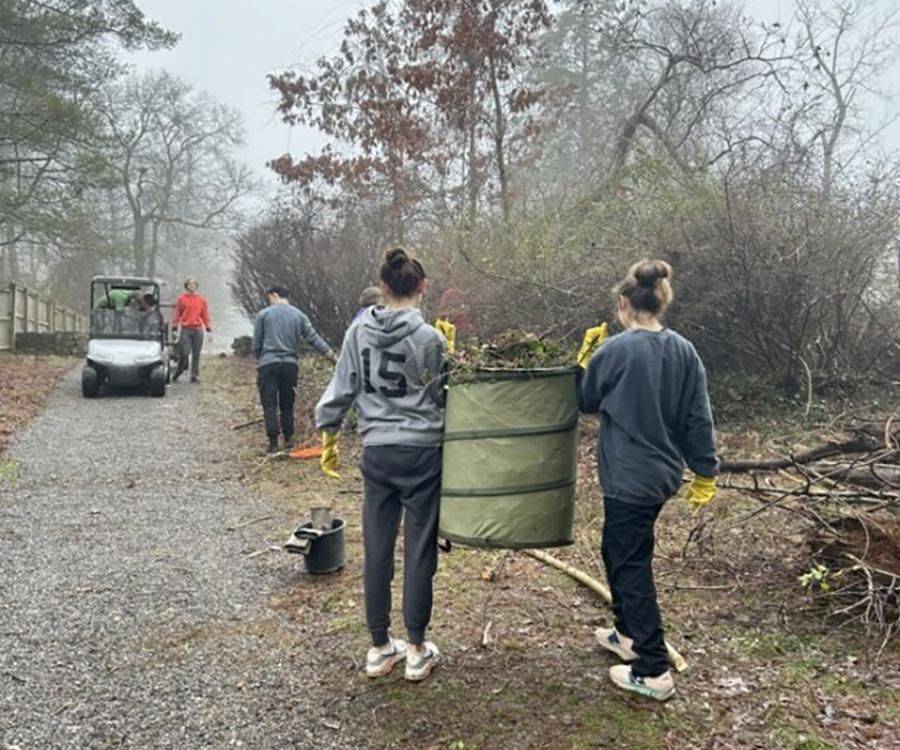 High school volunteers carrying a yard waste bag of pulled invasive plants along a gravel path during a weed wrangle at McCoy